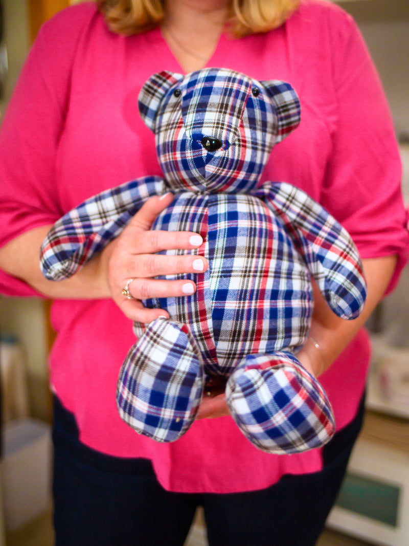 Woman wearing a pink shirt. Holding a stuffed bear made from a plaid shirt. It is a custom memory bear
