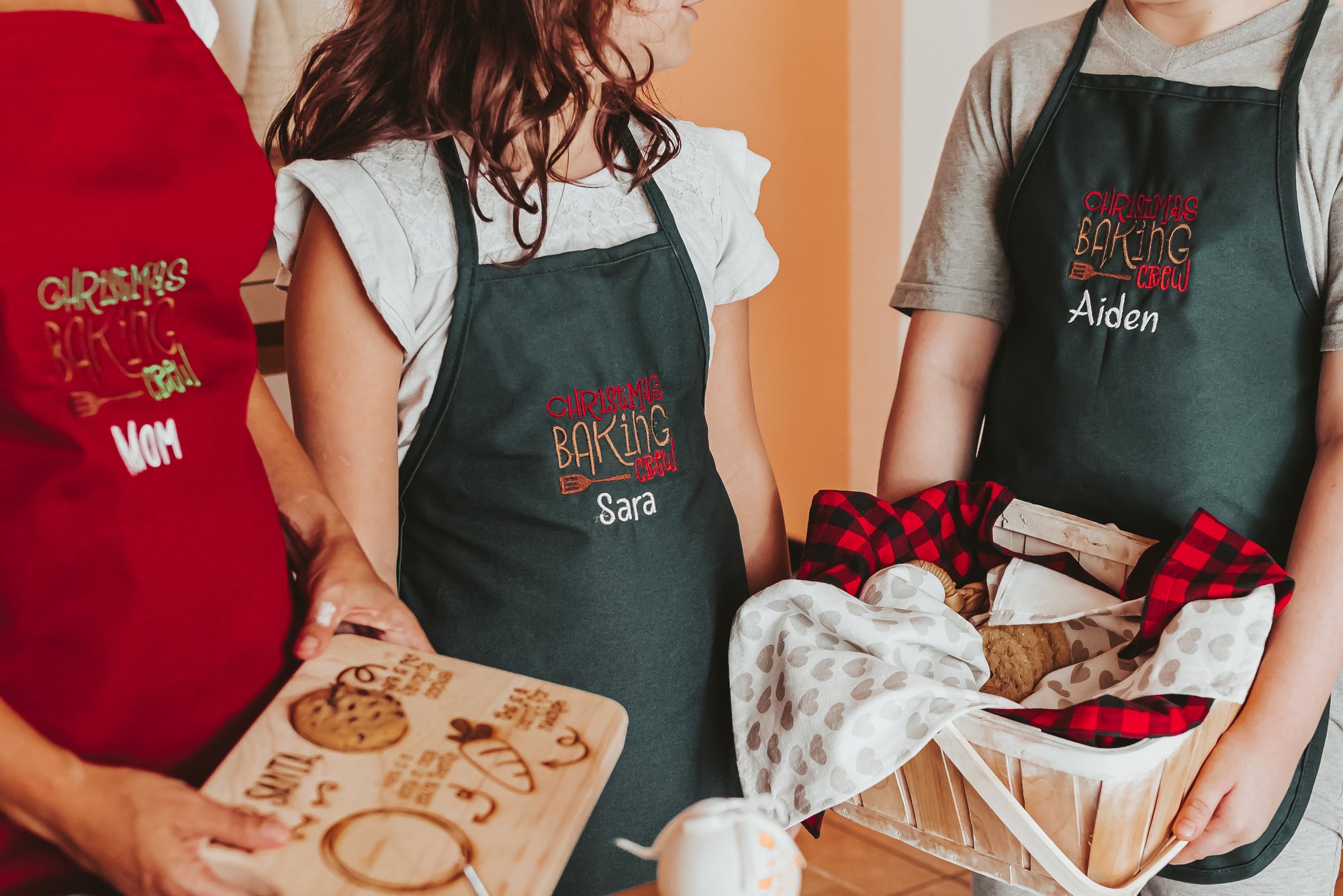 Mom and 2 children wearing aprons holding Christmas cookies. Aprons are machine embroidered with the phrase Christmas Cookie Crew. They are personalized with a name.