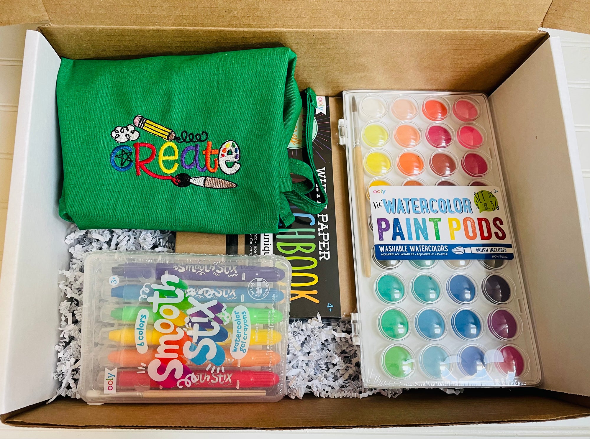 Green art smock featuring the word create embroidered on it. Has water colors, gel crayons and a sketch book.