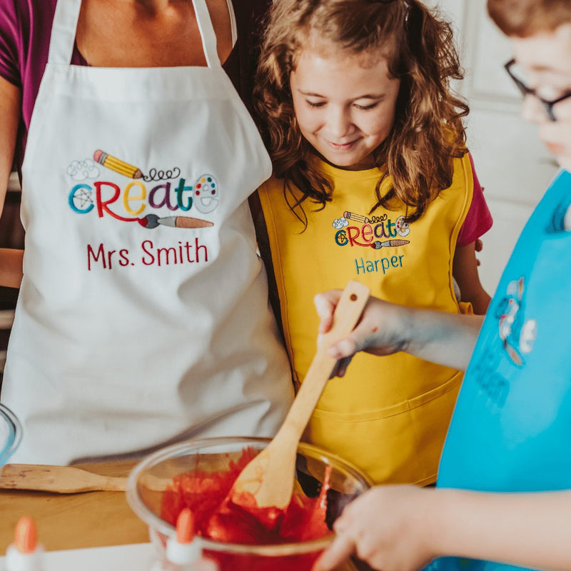 a mom wearing a white apron. a girl wearing a yellow smock and a boy wearing aturquiose smock. All embroidiered with the saying create. Also their name.