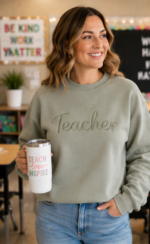 A smiling woman in a classroom setting wearing a sage green crewneck sweatshirt with the word 'Teacher' embroidered across the chest. She is holding a white travel tumbler with colorful text that reads 'TEACH love INSPIRE'.