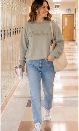 A woman walking down a school hallway wearing a sage green crewneck sweatshirt with 'Teacher' written across the chest, light blue jeans, white sneakers, and carrying a beige tote bag.