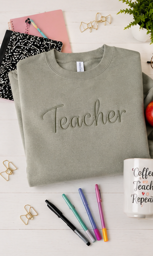 A folded sage green crewneck sweatshirt with the word 'Teacher' embroidered across the chest, styled in a flat lay on a white wooden background with notebooks, pens, binder clips, and a mug.