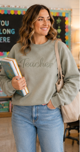 A woman wearing a sage green crewneck sweatshirt with 'Teacher' embroidered in gray script across the chest, paired with light blue jeans and carrying a beige tote bag.