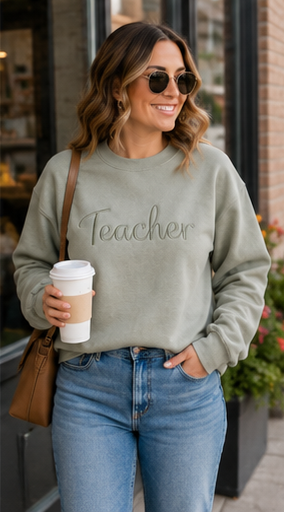 A woman wearing a sage green crewneck sweatshirt with the word 'Teacher' embroidered across the chest, paired with blue jeans, sunglasses, and a brown crossbody bag, holding a coffee cup in an outdoor setting.