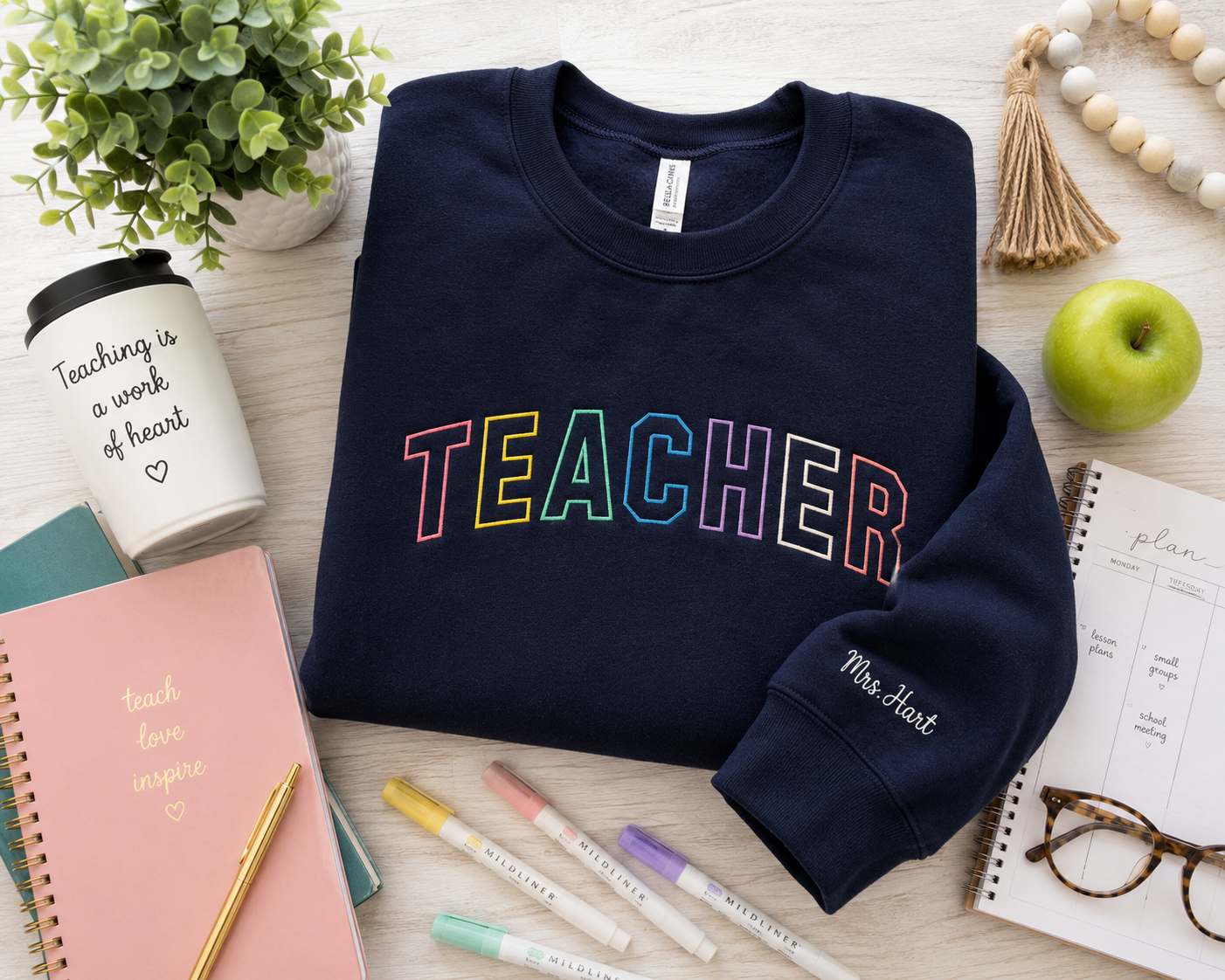 A navy blue crewneck sweatshirt with 'TEACHER' printed across the chest in rainbow-colored letters, styled in a flat lay with notebooks, pens, a plant, and other school-themed props on a light wood background.