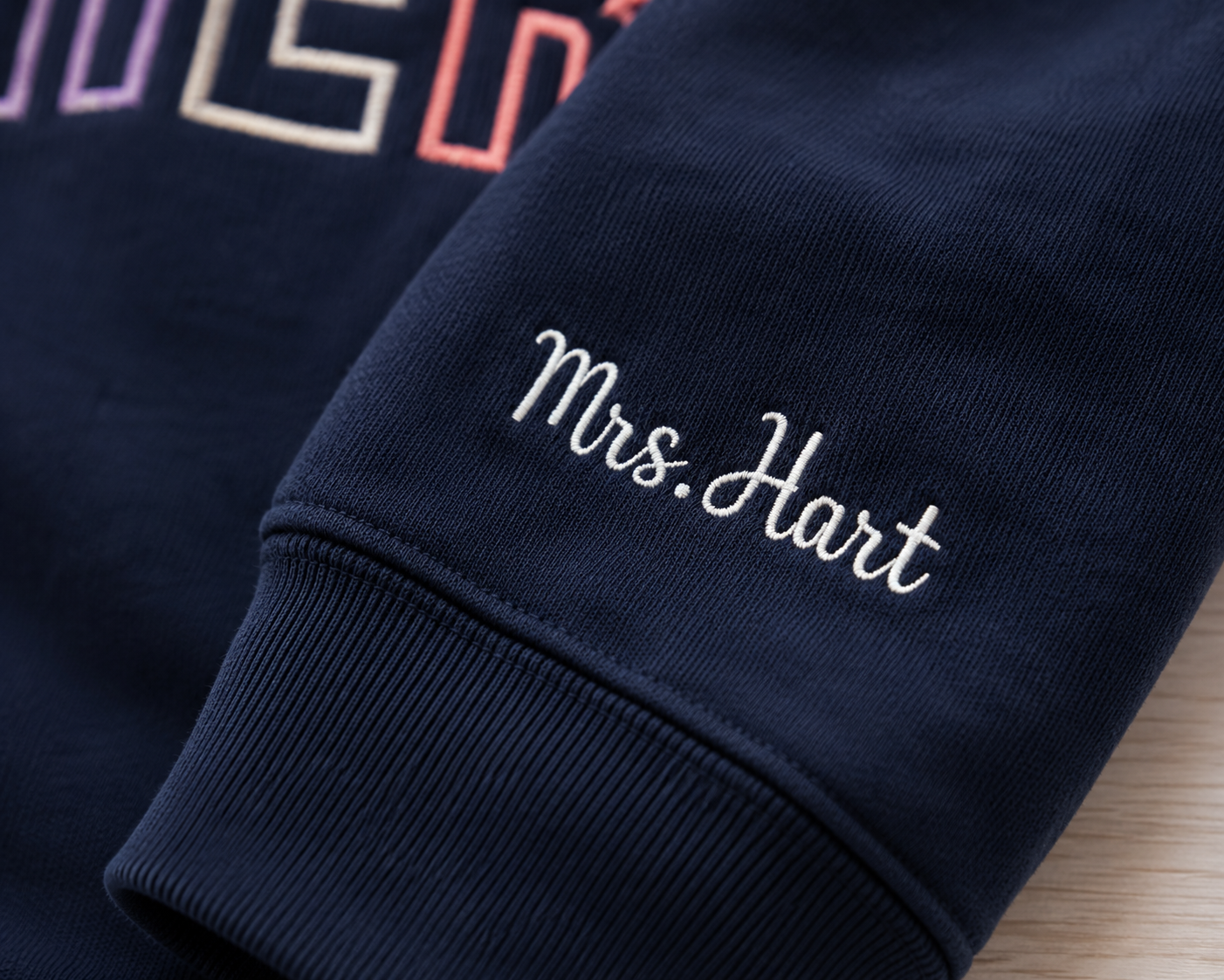 Close-up of a navy blue sweatshirt cuff with white embroidered text reading 'Mrs. Hart' and part of a multicolor graphic at the top.
